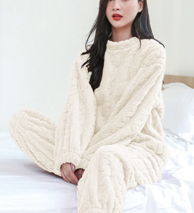 Cozy woman in a cream Winter Coral Velvet Pajama Set with a pullover and pants, relaxing on a bed by PureAura.