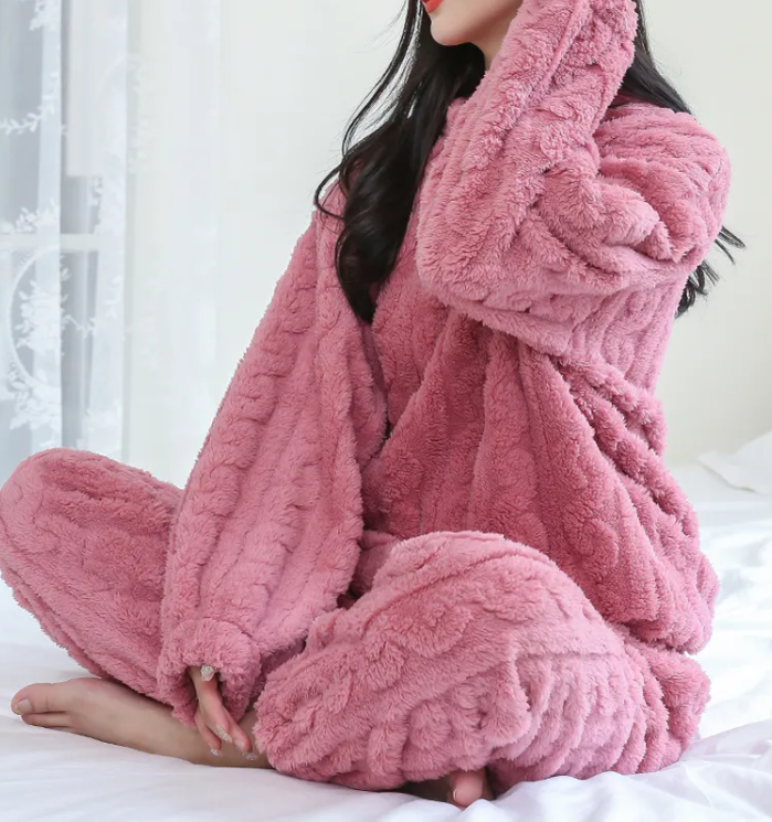 Cozy woman wearing a pink Winter Coral Velvet Pajama Set – Warm Pullover & Pants by PureAura, sitting on a bed.