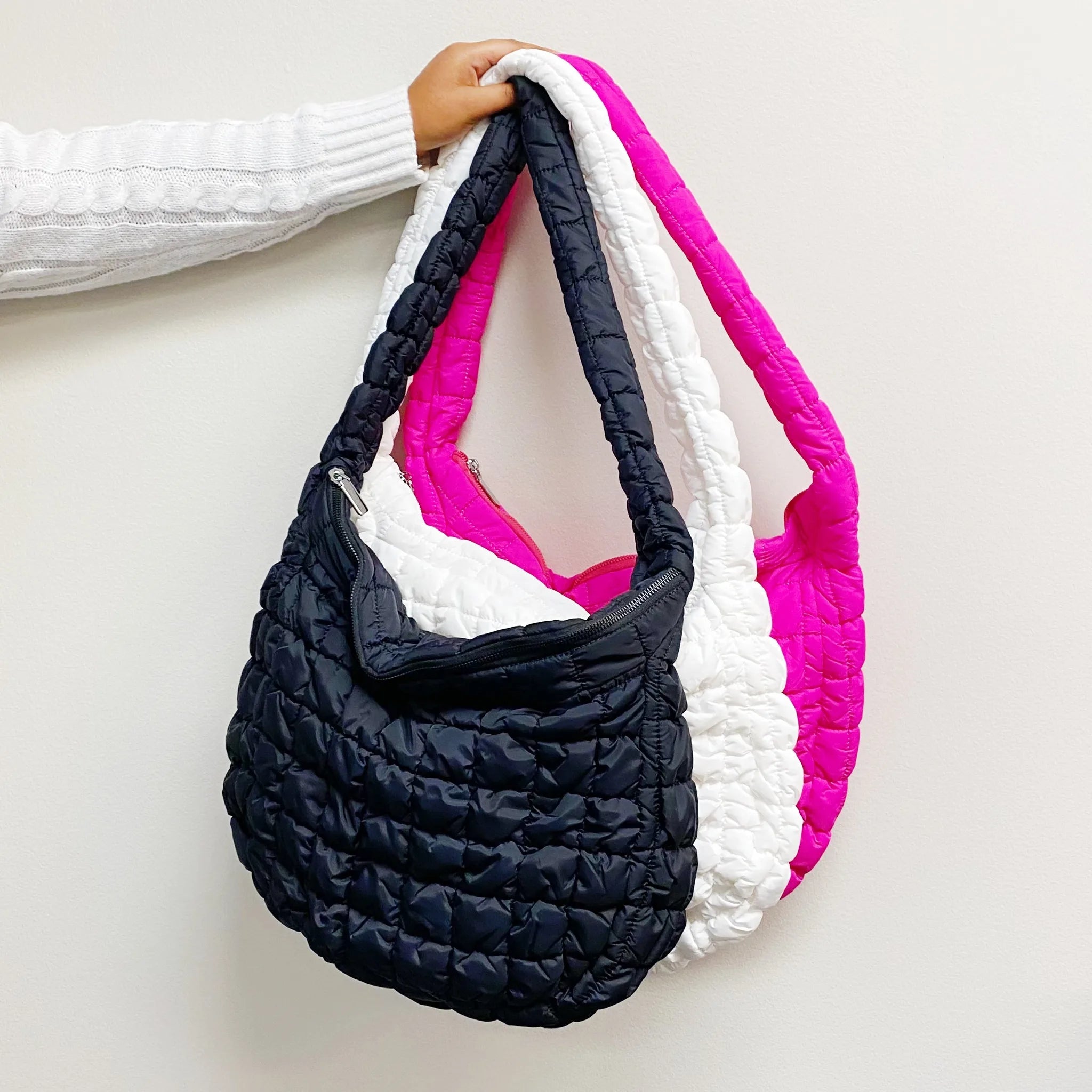 Three colorful Carry All Quilted Cross Body Bags in black, white, and pink, held by a person against a white wall.
